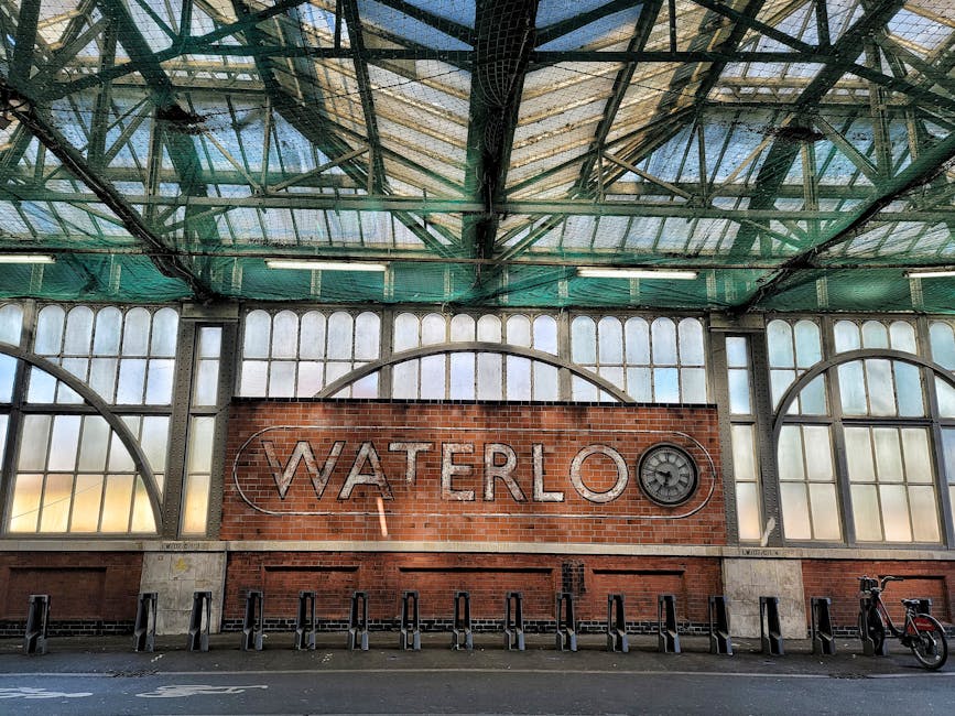 The exterior of a large, modern train station building with a distinctive curved glass facade and a prominent red railway symbol centered above the entrance. The lower part of the building features a 